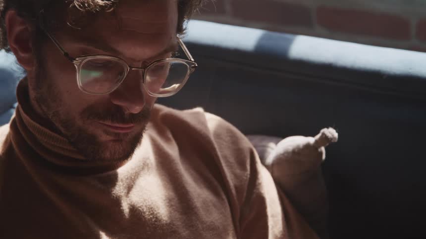 Young bearded man wearing glasses sits in a couch in a room reading