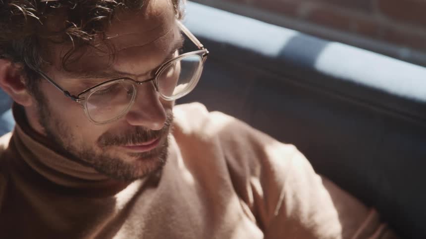 Man with glasses a beard and a brown turtleneck sweater reads a book on a sofa
