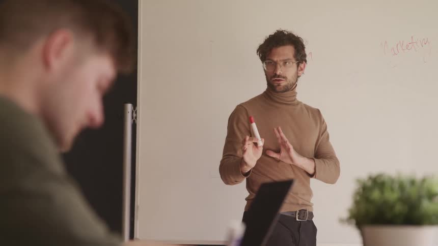 A man standing and talking holding a red marker with a whiteboard behind him