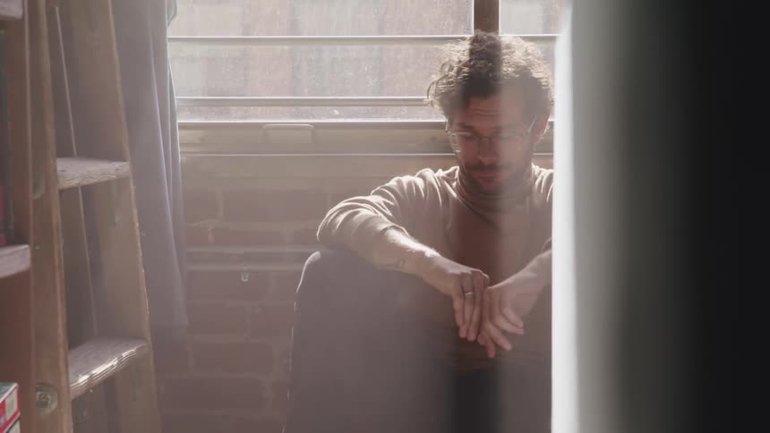 Man with a brown turtleneck sweater glasses and a beard sits on the ground and leans against a wall under a window in the sunlight in a room