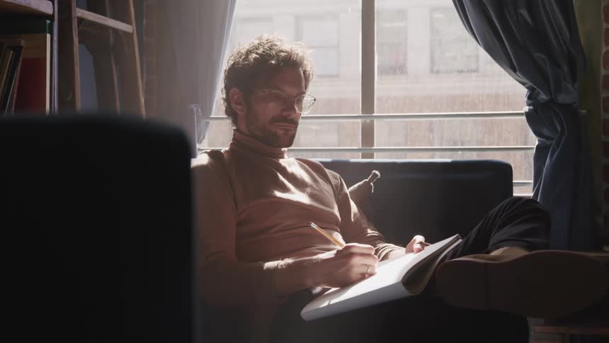 A man sits on a small couch in the corner of a study room with a pencil and notebook