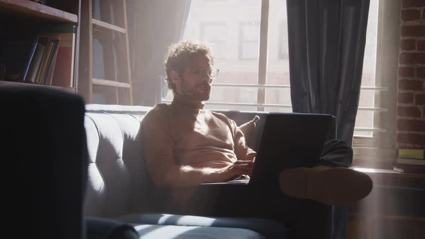 Man with a beard glasses and a brown turtleneck sweater sits on a couch and uses a laptop in a room