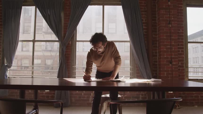 A man with a pencil writing on a paper on a table with windows in the background