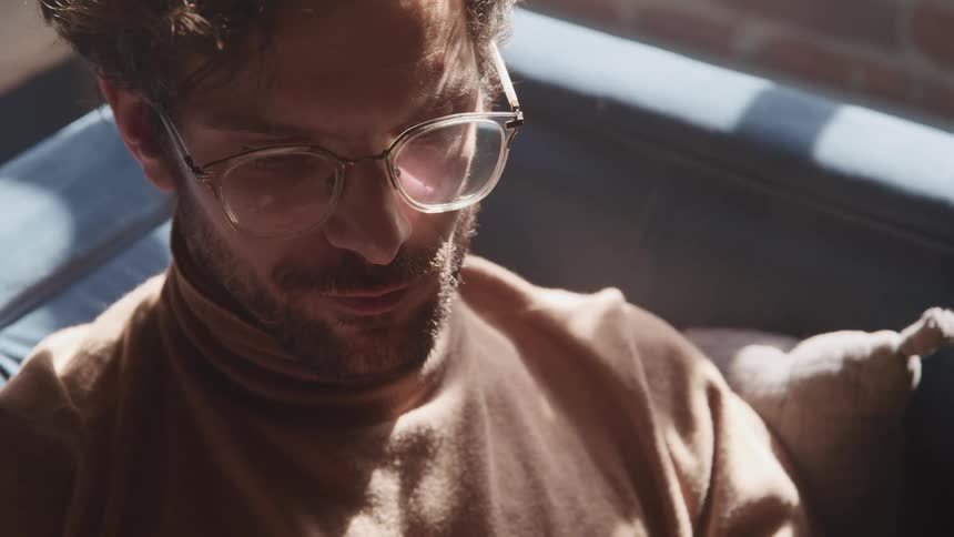 Man with a glasses a beard and a brown turtleneck sweater sits on a couch and reads a book