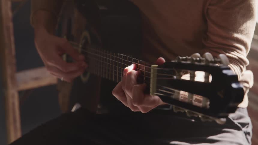 Man with brown pants and a sweater sits on a stool and plays a guitar