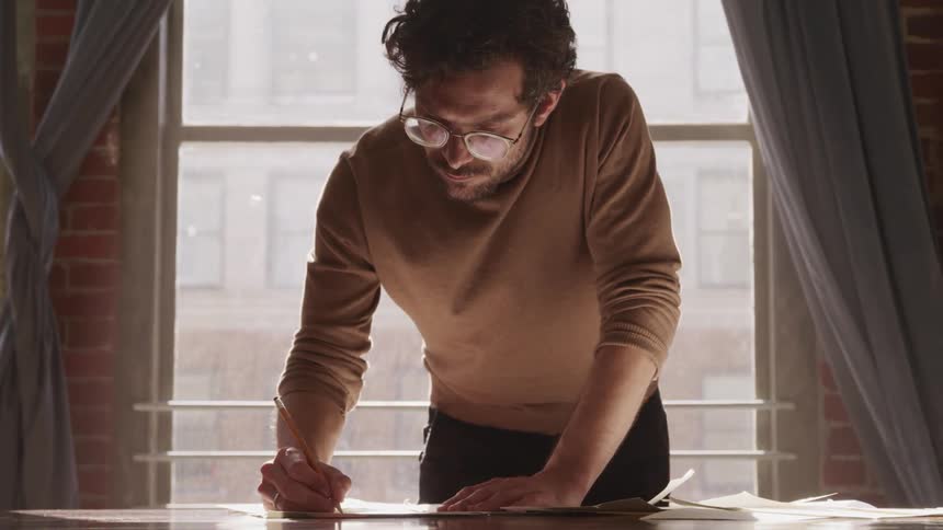Man leans on a desk in front of a window and draws on paper with a pencil
