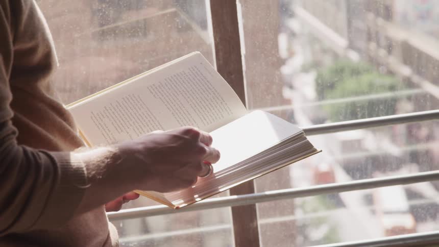 A man reads a book by a window and then quickly rips a page out and crumples it up