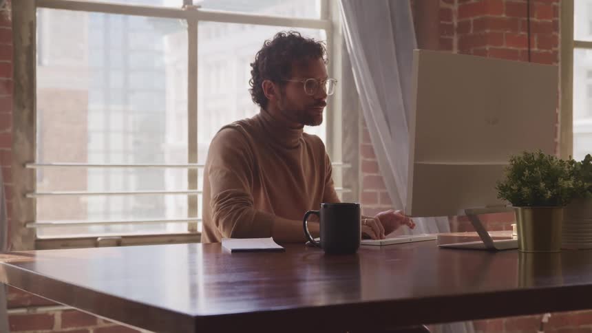 Bearded man is working on computer at desk in front of a large window set in brick walls