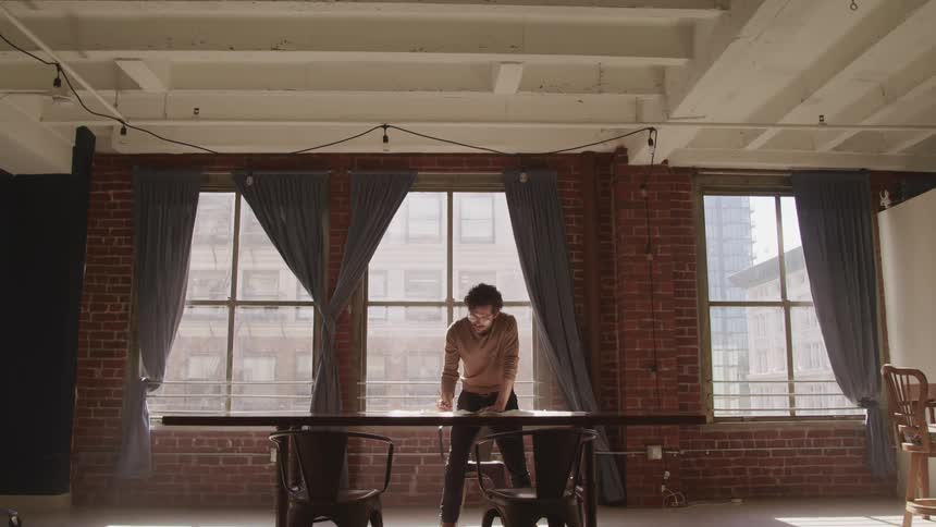 Man stands looking down on papers on table near three big windows