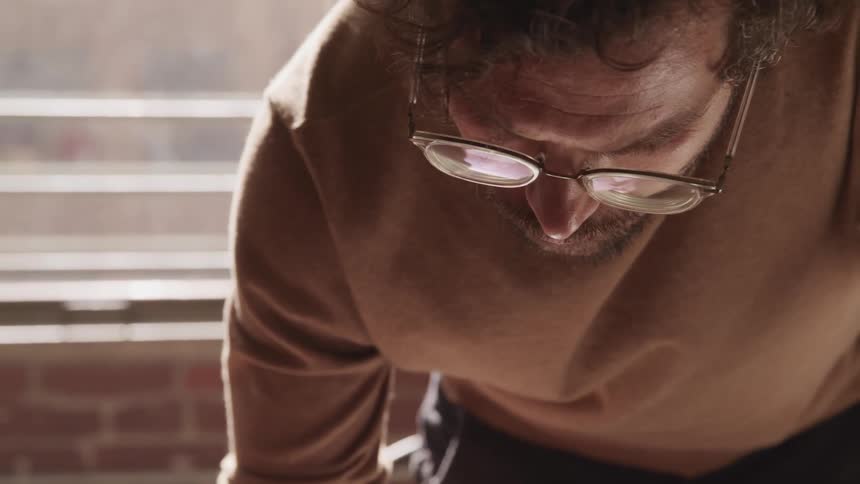Man wearing glasses bends over at the waist while near a window