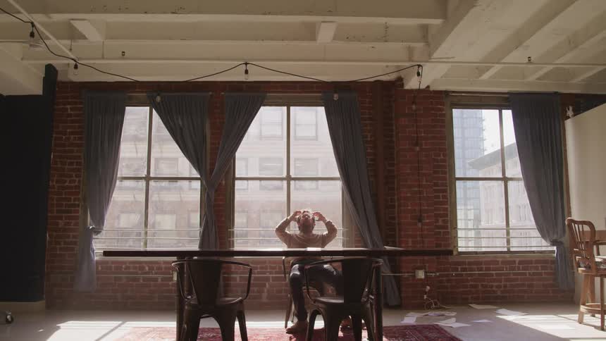 A man sits in his desk chair in his apartment and runs his hands through his hair as leans back