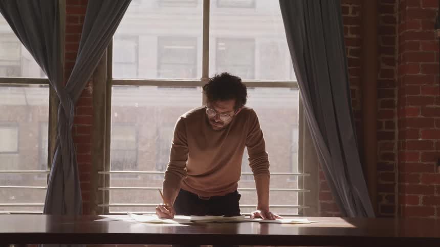 A man stands at his desk in front of a large window and writes on papers