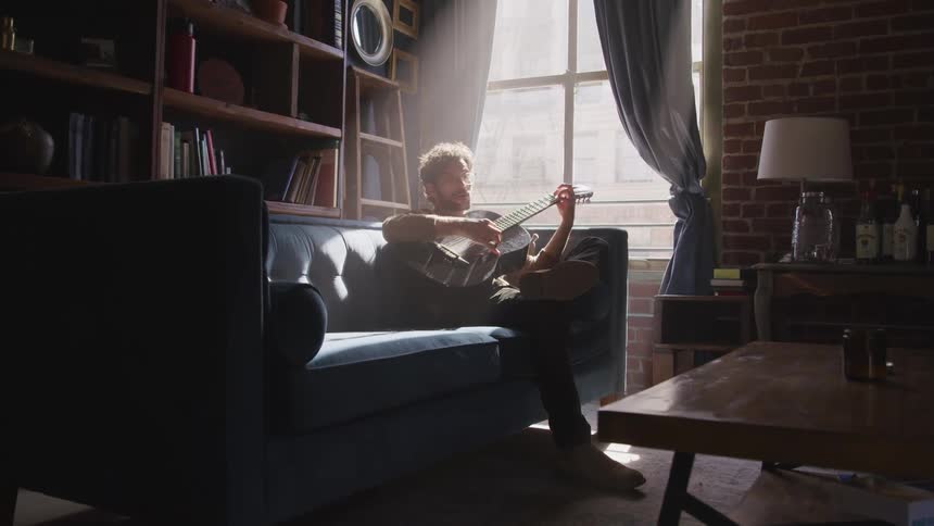 A male caucasian sitting on a couch playing a guitar