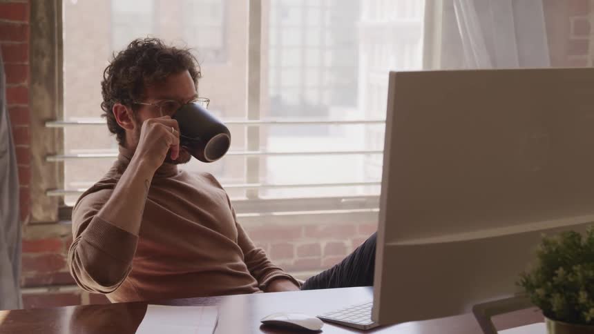 Young man wearing turtleneck and glasses sits at the office in front of a computer while drinking a hot beverage