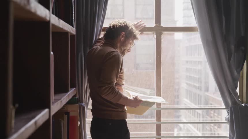 A man looks out his window and reads a hardcover book while standing next to a large shelving unit