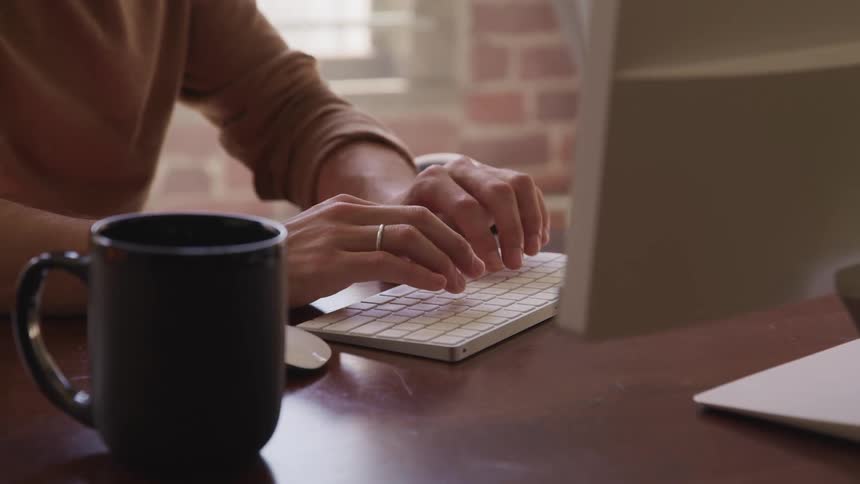 Man with a brown sweater sits at table and types on the keyboard in a room