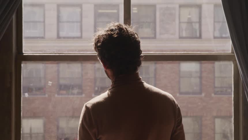 Man with a brown turtleneck sweater looks out the apartment window