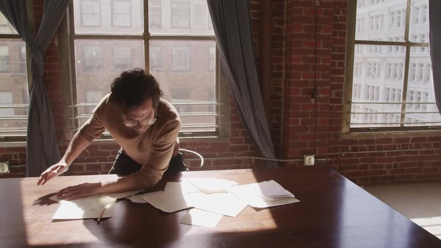 Man studying papers on table in room with view of city abruptly tosses them to the floor