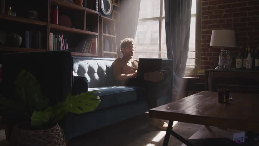 Man with a beard glasses and a brown turtleneck sweater sits on a sofa and uses a laptop as the sun shines through windows in the living room