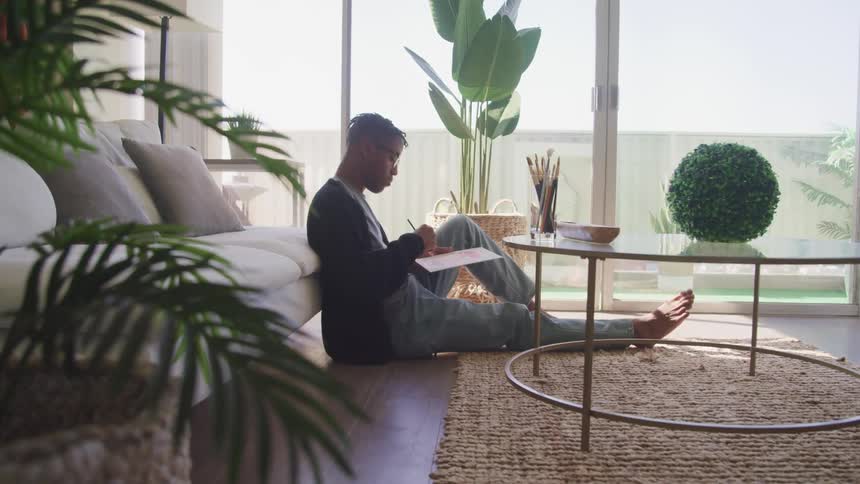 A man sits on the floor in front of his couch casually painting or writing on a board