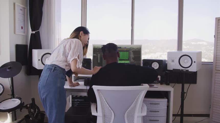 A man sits in front of a computer using audio software as a young asian woman stands next him with her hand on his shoulder