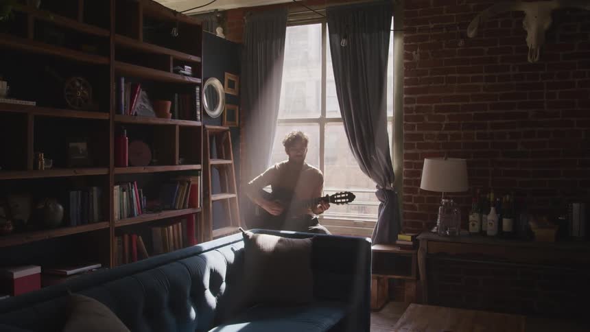 Musician plays the guitar while sitting next to the window in the living room under the sunlight