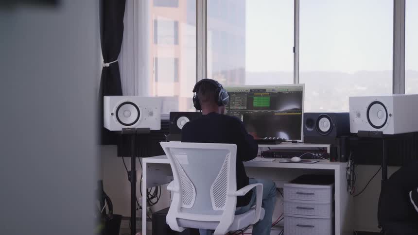 A person sitting in front of a desk with a computer and speakers with a window behind it