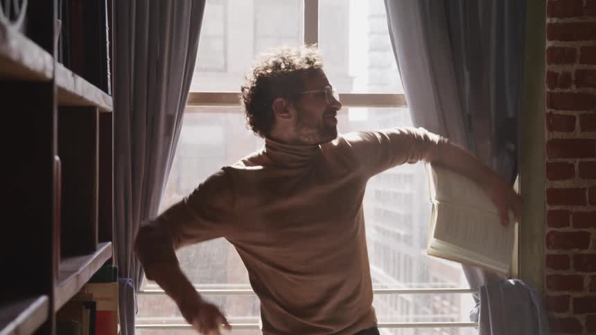A man standing next to a large window tears a page out of his notebook and throws it in frustration