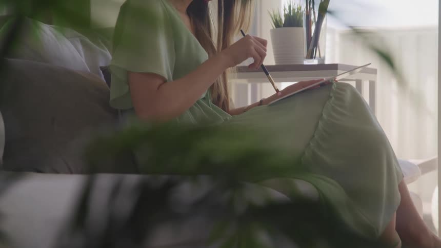 A woman sits on a gray couch and paints on a canvas on her lap