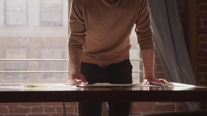 Man aggressively sweeps documents from a table in a room in front of a window