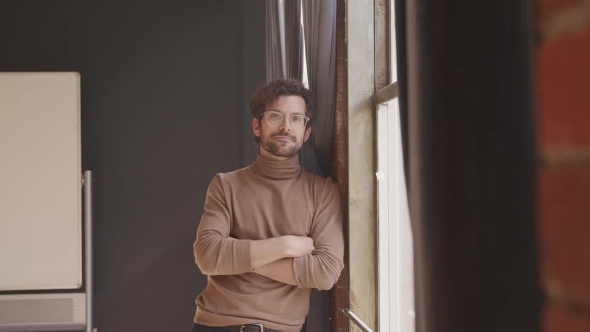 Man with a beard a turtleneck sweater and glasses stands next to a window and faces the camera with eye contact