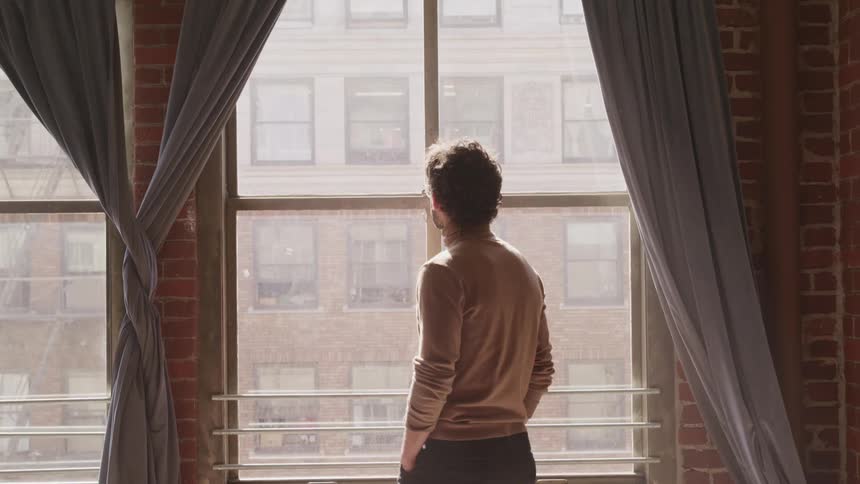 Man with a brown turtleneck sweater a beard and glasses walks and looks out the apartment window