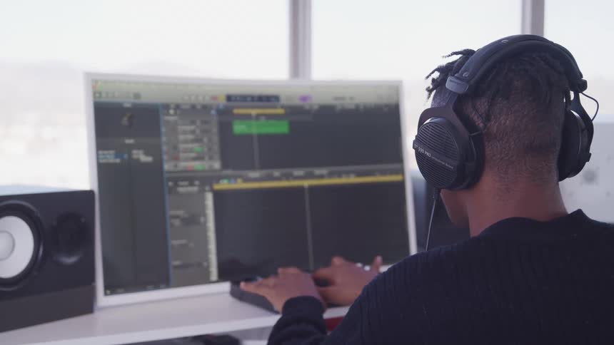 Man wearing headphones sits at table near window manipulating keyboard of computer bordered by speakers