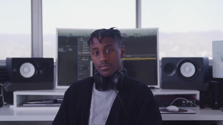 A young man with headphones around his neck and sitting with his back to a desk holding a computer and large speakers turns his head to look into the camera