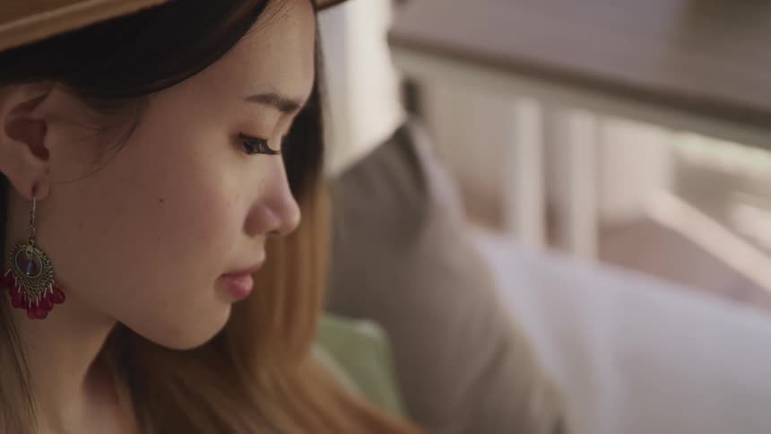 Asian woman with a brown hat earrings and makeup sits on a sofa and looks down in a room