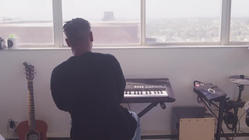 A man sits at a keyboard with other instruments around him in front of a highrise window