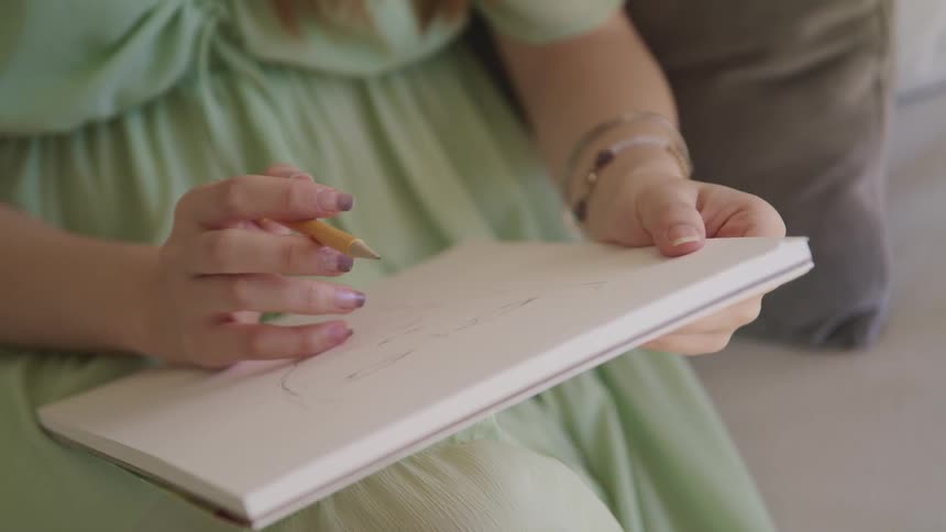 Woman in a green dress draws with pencil on paper while sitting in a room