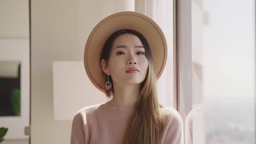 Asian woman with beige hat and a sweater looks out the window and turns her gaze to the camera in a room