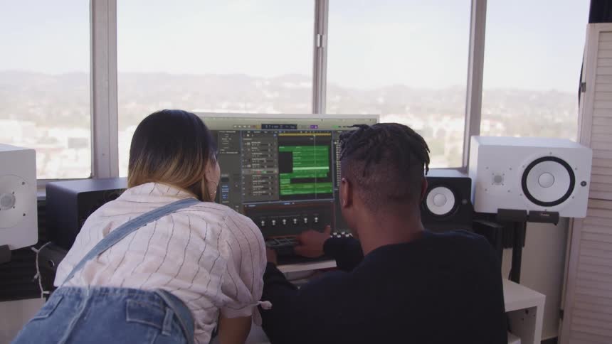 Woman leans over to look at screen as man manipulates settings at desk bordered by speakers