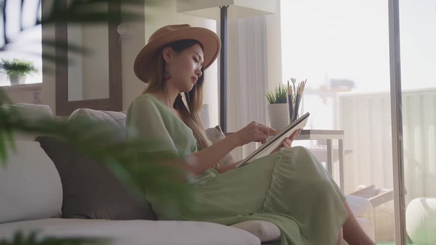 A young asian woman sits on a sofa in a house while drawing on a notepad with a pencil