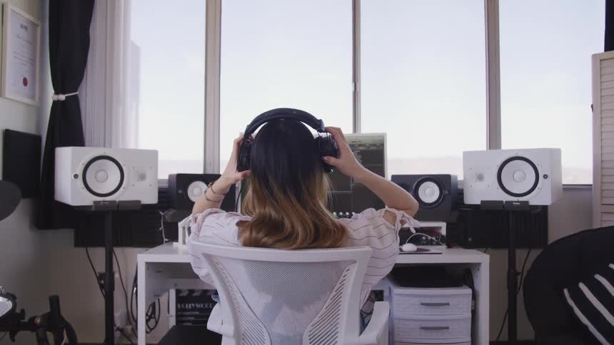 A woman sitting at a desk putting on headphones in front of a computer and speakers with a window in the background