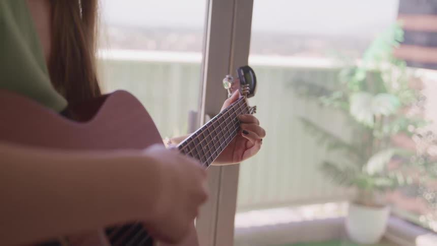 A woman plays an acoustic guitar next to windows