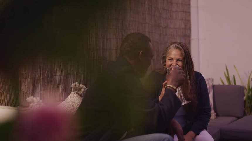 A man and woman sit on a couch and laugh as they each drink a glass of wine