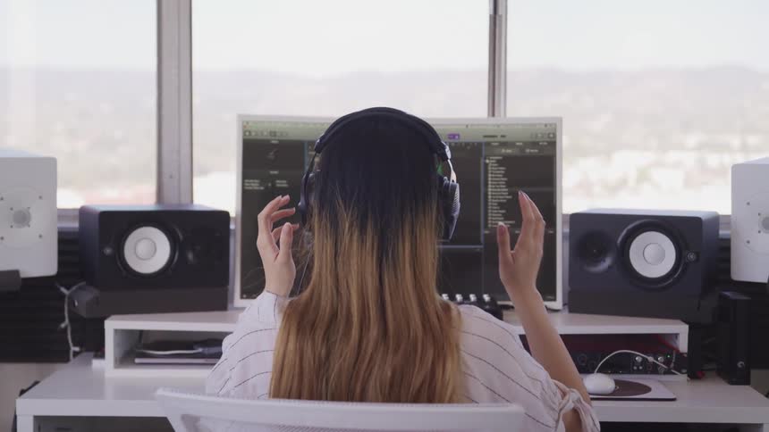A woman sits in her office surrounded by speakers and puts on headphones and starts to use her computer