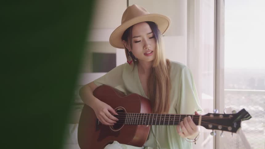 Woman plays guitar sitting near glass doors showing balcony
