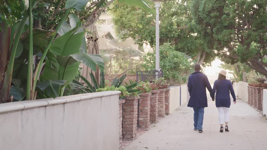 A man and woman hold hands as they walk down a sidewalk in a tree lined neighborhood