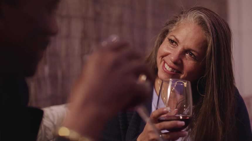 Woman with grey hair and hoop earrings drink a glass of wine with black man