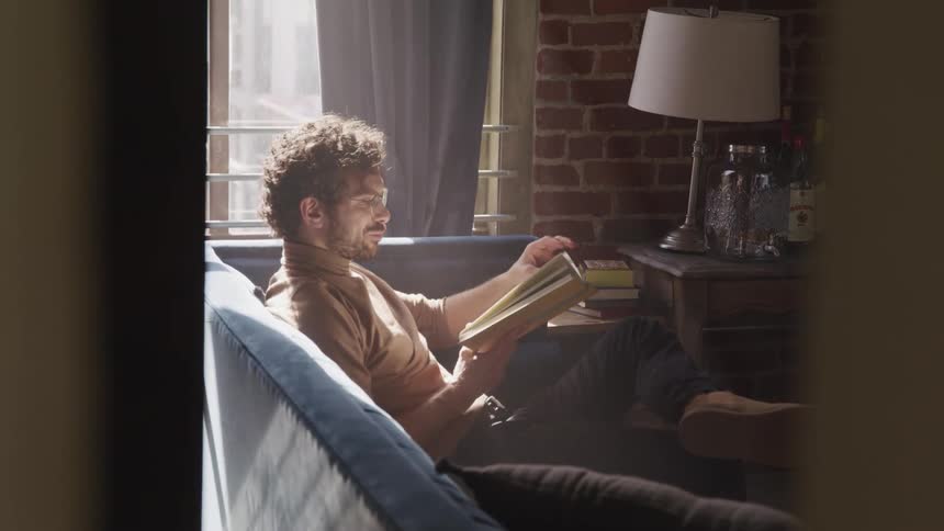 A man sits on a couch in the corner of a room reading a book