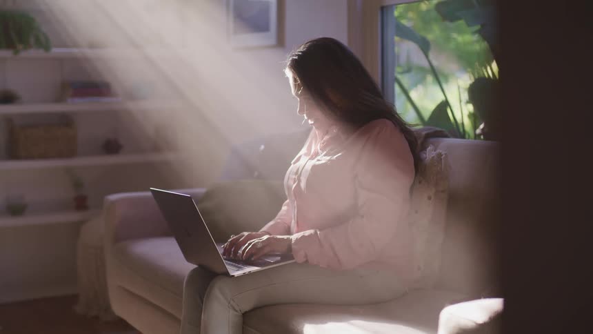 A woman sits at home with her laptop in her lap and types