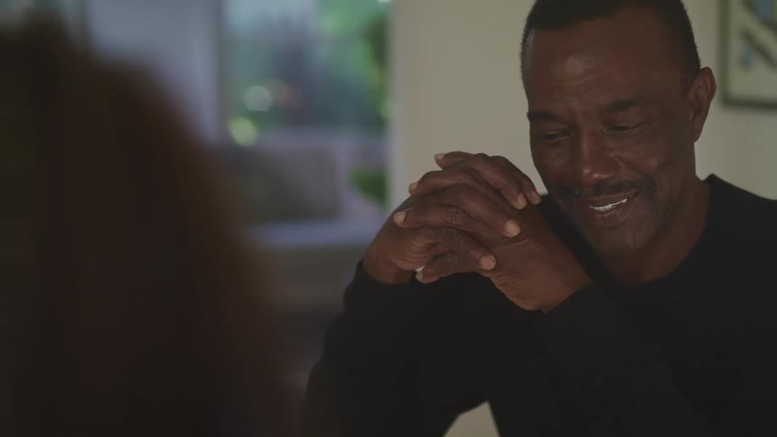 A man at home smiles as he sits across from another person